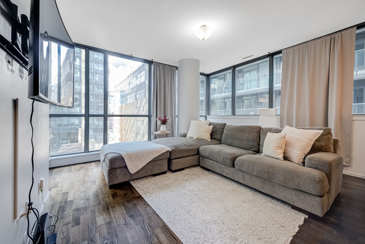 A spacious living area features large windows allowing natural light to fill the room, with soft drapes providing privacy. A comfortable sectional sofa sits on a light area rug, complemented by a throw blanket and accent pillows. A wall-mounted television is visible, enhancing the entertainment options.