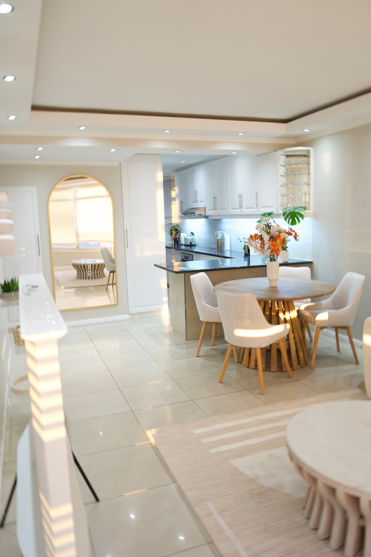A modern kitchen and dining area feature sleek cabinetry and a circular dining table surrounded by light-colored chairs. Natural light brightens the space, reflecting off polished tiles. An archway leads to a living area, which includes a textured rug and decorative plants.