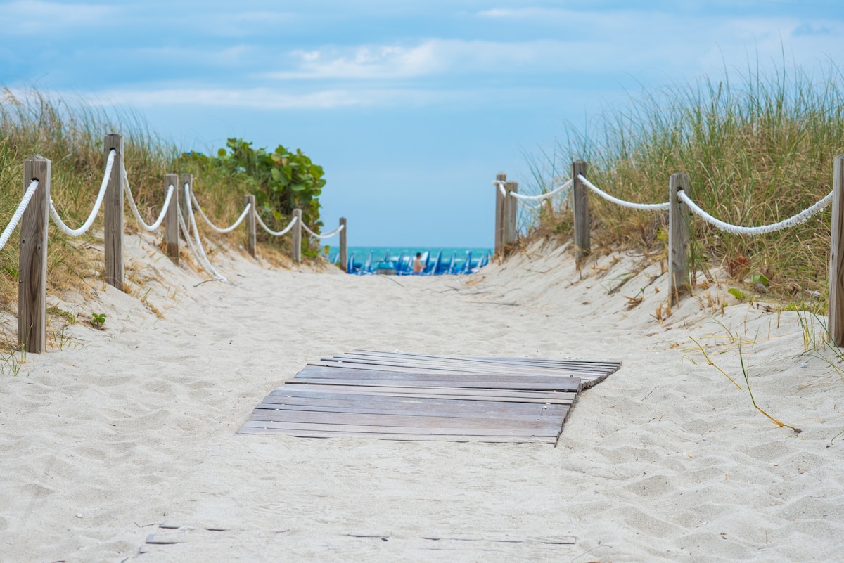 A wooden pathway leads through soft, sandy terrain bordered by tall grasses and shrubs. The pathway directs towards a picturesque view of the ocean in the distance, with hints of blue water visible beyond the dunes.