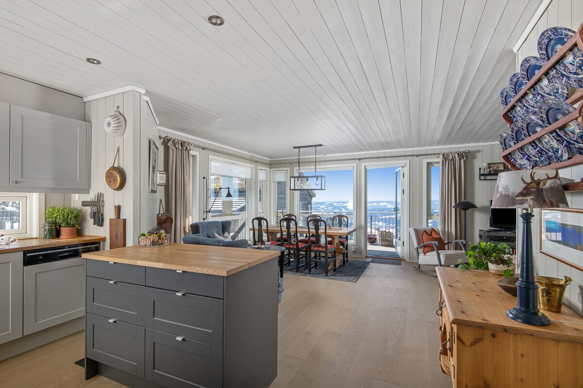 An open-plan living area is showcased with a large kitchen island at the center. Windows provide scenic views and natural light, complemented by a dining table and chairs arranged nearby. Decorated shelves display ceramic dishes, adding character to the spacious design.