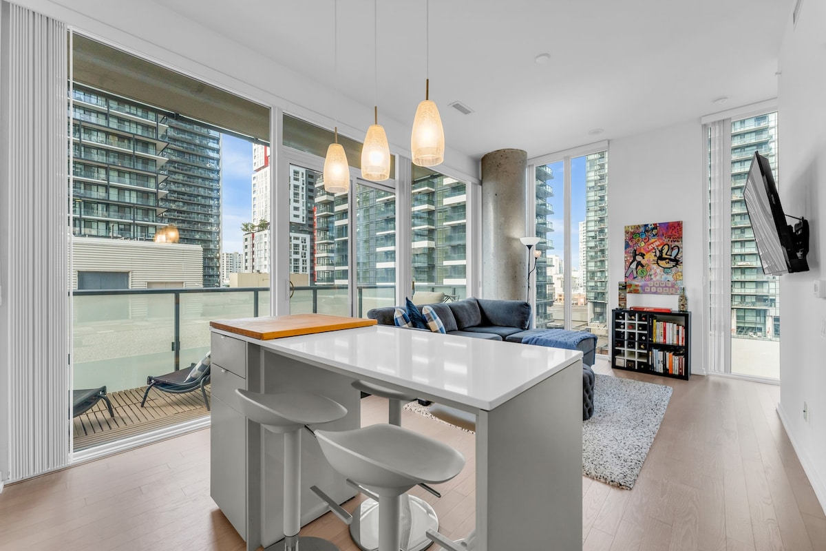 The open living area is illuminated by natural light from floor-to-ceiling windows. A modern kitchen island with seating is positioned near the living space, complemented by a comfortable sofa and a small coffee table. Decorative elements add a vibrant touch to the room.