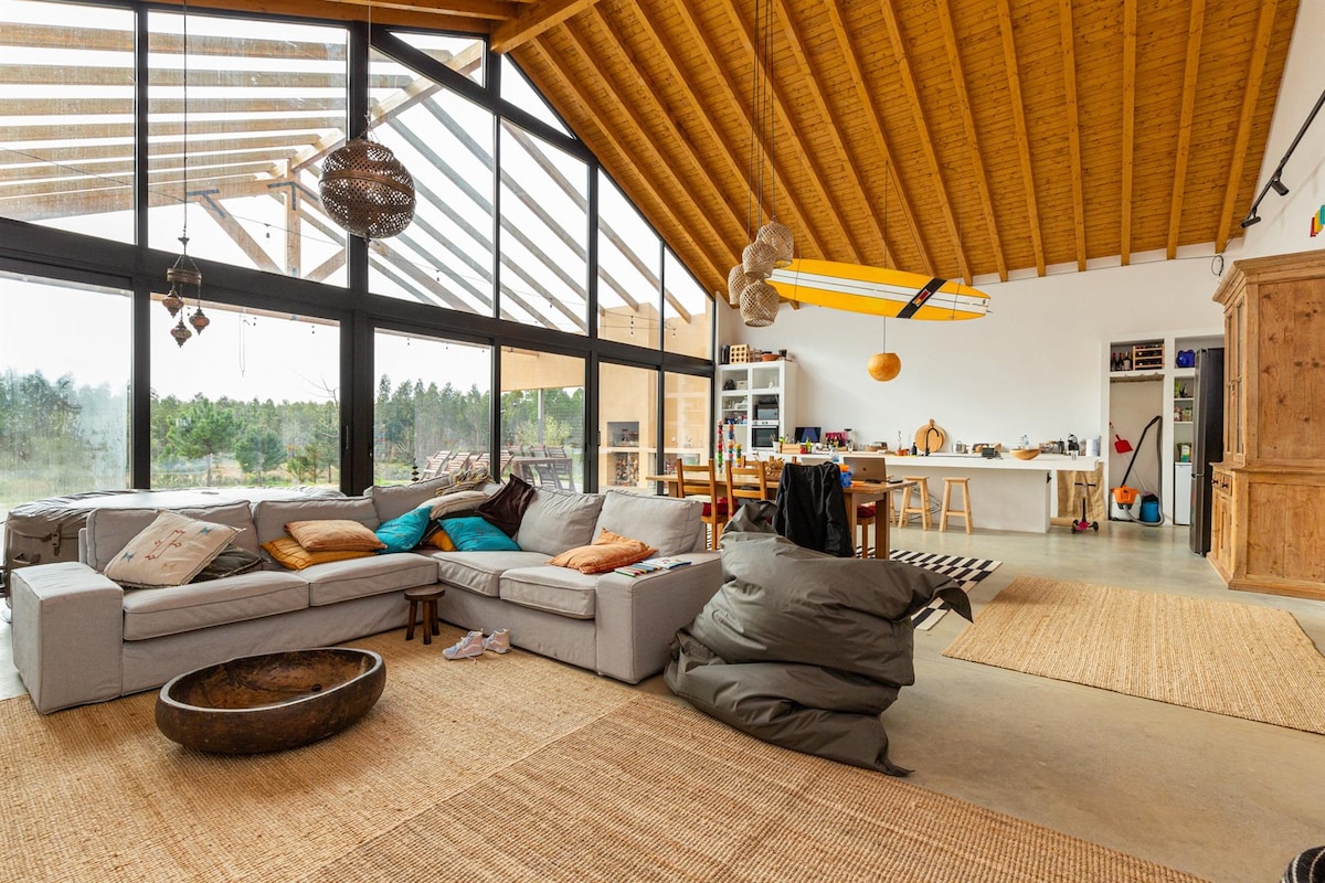 A spacious living area features a large sectional sofa adorned with colorful cushions and multiple bean bags scattered on a natural fiber rug. Large windows bring in ample light, with a dining area and kitchen visible in the background, showcasing a modern, open layout.