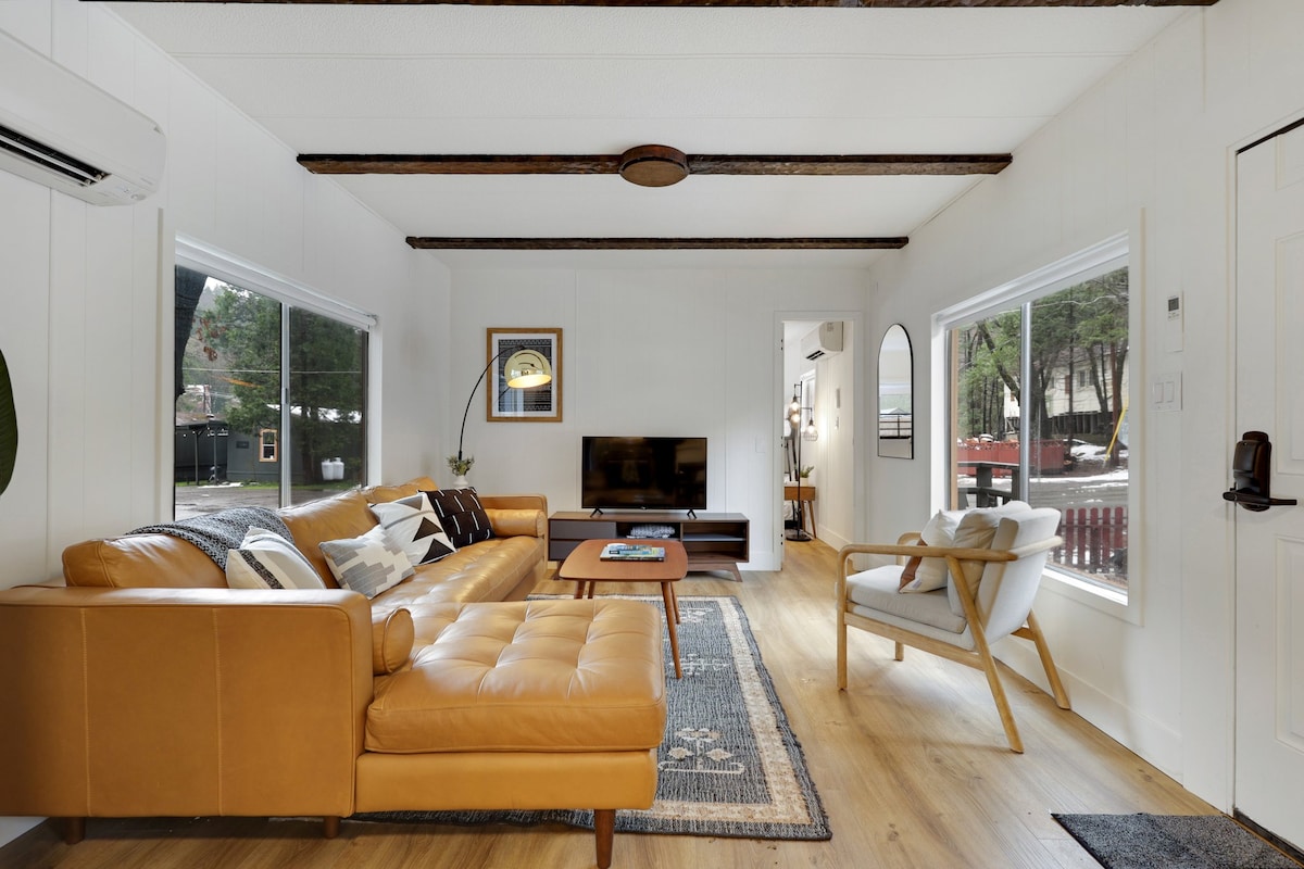 A modern living area features a leather sectional sofa, paired with an elegant armchair. Natural light filters through large windows, while a wooden coffee table sits on a decorative area rug. A wall-mounted television offers entertainment options, complementing the overall inviting space.
