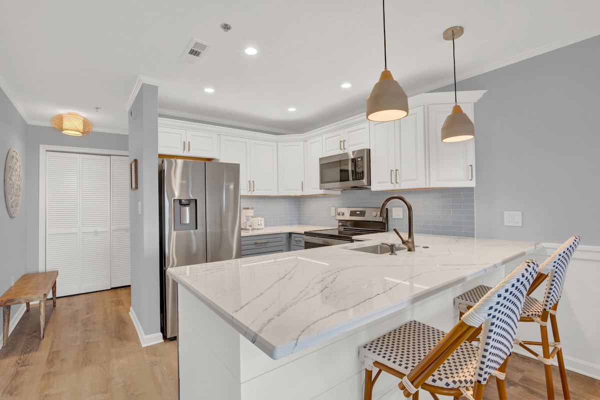 A modern kitchen features a spacious island with a marble countertop, equipped with high-backed chairs. Stainless steel appliances and bright white cabinets contrast with soft blue accents on the backsplash, creating a clean and functional cooking space. Ample lighting enhances the open layout.