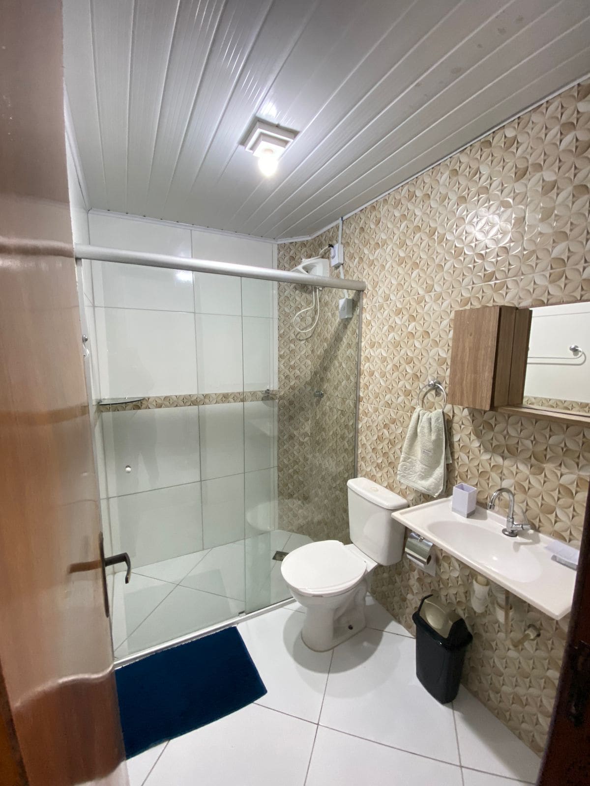 A well-lit bathroom features modern beige and white tile walls. A glass shower enclosure is visible alongside a compact toilet and sink. A large mirror above the sink enhances the sense of space, and a small wall cabinet offers additional storage.