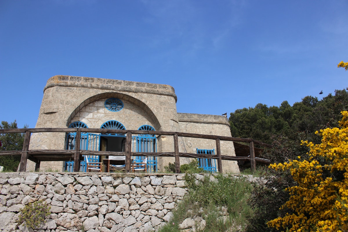 A stone structure stands on elevated terrain, featuring blue-framed windows and a circular window above the entrance. A wooden railing surrounds the patio, where rustic chairs are positioned. Vibrant yellow flora grows nearby, contrasting with the greenery and clear blue sky.