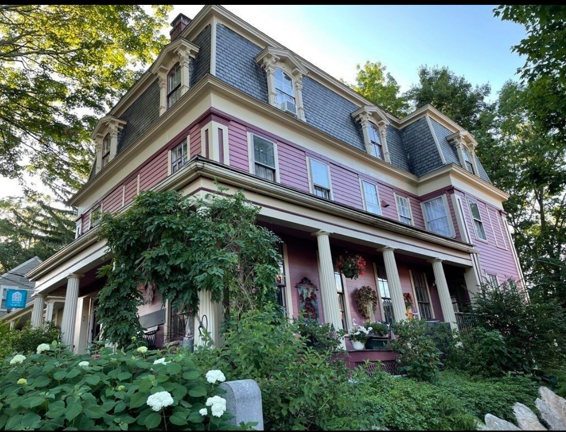The exterior of a historic building features a striking pink façade with decorative trim and large windows. Lush greenery surrounds the structure, complemented by blooming flowers and hanging baskets. A welcoming porch is supported by tall columns, enhancing the building's grand appearance.