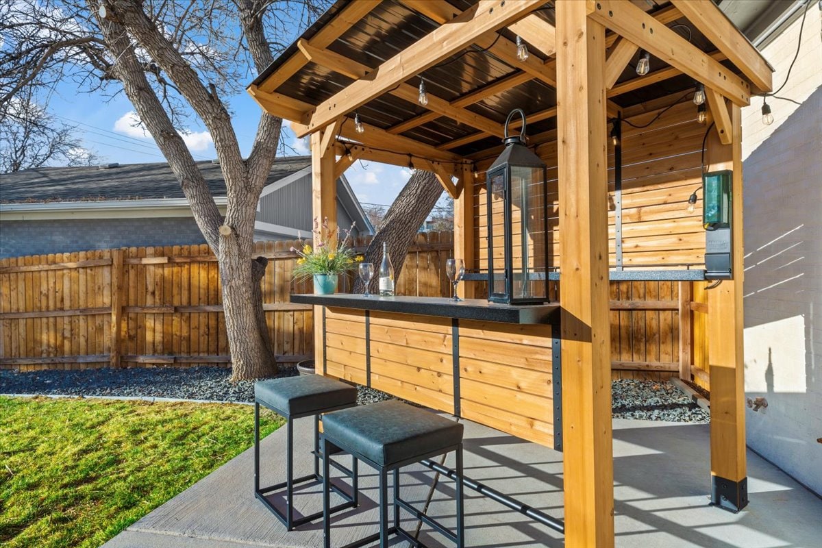 An outdoor bar structure is featured, constructed with warm wooden beams and a dark countertop. Two stylish stools are positioned beneath. Soft lighting is provided by hanging fixtures, while a lush green lawn and wooden fence create a private setting.