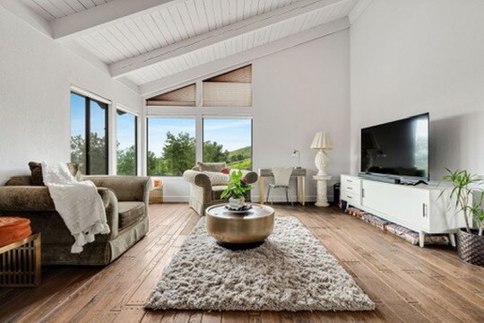 A spacious living area is highlighted by large windows offering views of the surrounding landscape. Neutral furnishings include a cozy sofa and armchair, arranged around a central coffee table on a textured rug. Natural light fills the room, enhancing the warm wooden flooring.