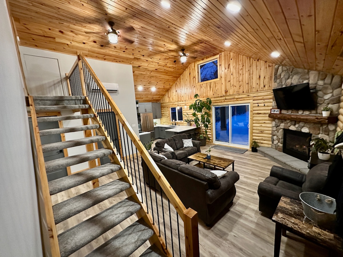 An inviting living space with high wood-paneled ceilings and large windows allowing natural light. Cozy sofas are arranged around a stone fireplace, complemented by a wooden coffee table. A staircase leads to an upper level, enhancing the openness of the area.