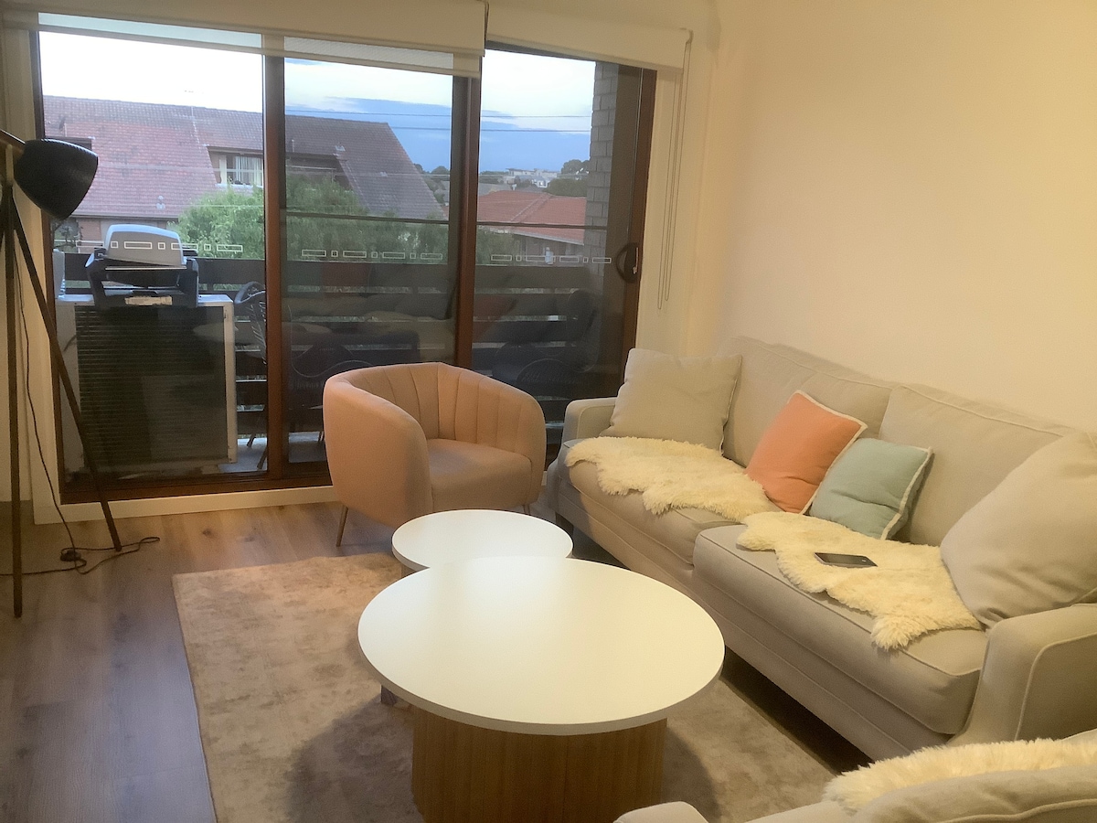 A cozy living area is furnished with a light-colored sofa and a stylish armchair. A round coffee table sits in the center, accompanied by a soft area rug. Large windows offer a view of the outside, allowing natural light to fill the space.