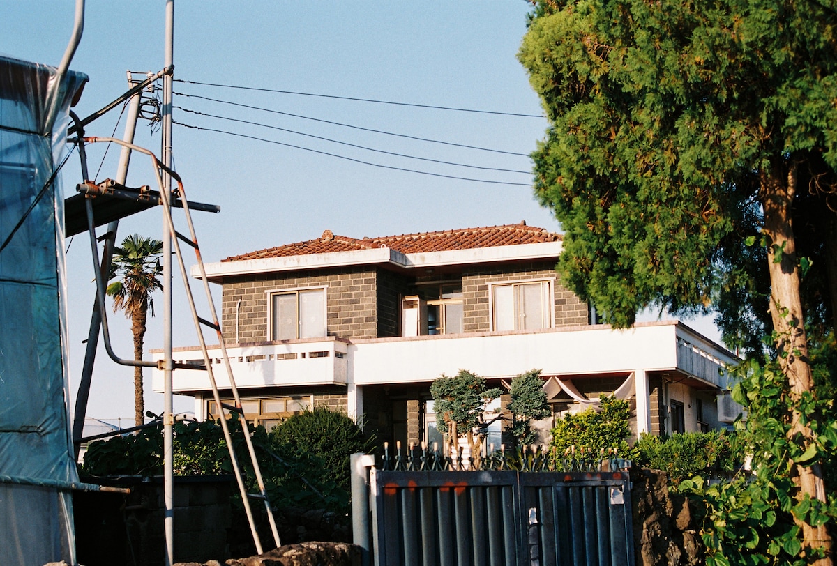 A two-story building is set against a clear blue sky, showcasing traditional architecture with a red-tiled roof. Surrounding greenery includes a tall tree and various plants. In the foreground, a fence and scaffolding are visible, adding to the farm-like scenery.