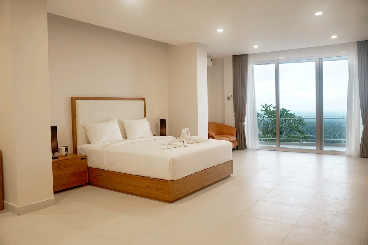 A spacious bedroom features a large bed with neatly arranged linens, accented by wooden bedside tables. Large glass doors lead to a balcony, providing panoramic views of the surroundings. Natural light floods the room, complemented by light-colored walls and simple decor.
