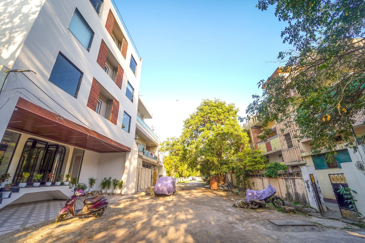 A quiet street view is captured, showcasing low-rise buildings flanking each side. Trees provide shade, and two parked vehicles are visible on the unpaved road, surrounded by greenery. Natural light creates a welcoming atmosphere in this neighborhood setting.