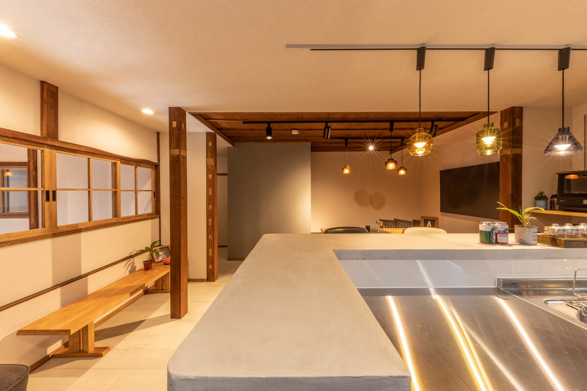 The spacious kitchen features a central concrete island with a stainless steel countertop, surrounded by contemporary lighting fixtures. Wooden beams and a bench add warmth, while the open layout invites gatherings. A dining area is visible in the background, enhancing the overall communal space.