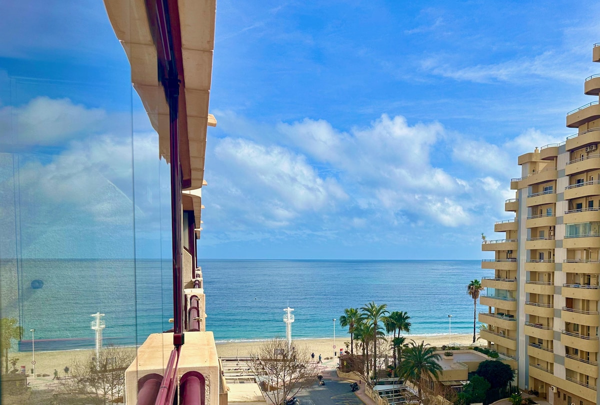 A view from the sixth floor showcases the beach and sea, framed by modern buildings. The sandy shoreline extends into the distance, with gentle waves meeting the shore. Palm trees line the beach, and a clear blue sky with scattered clouds completes the serene ocean scene.