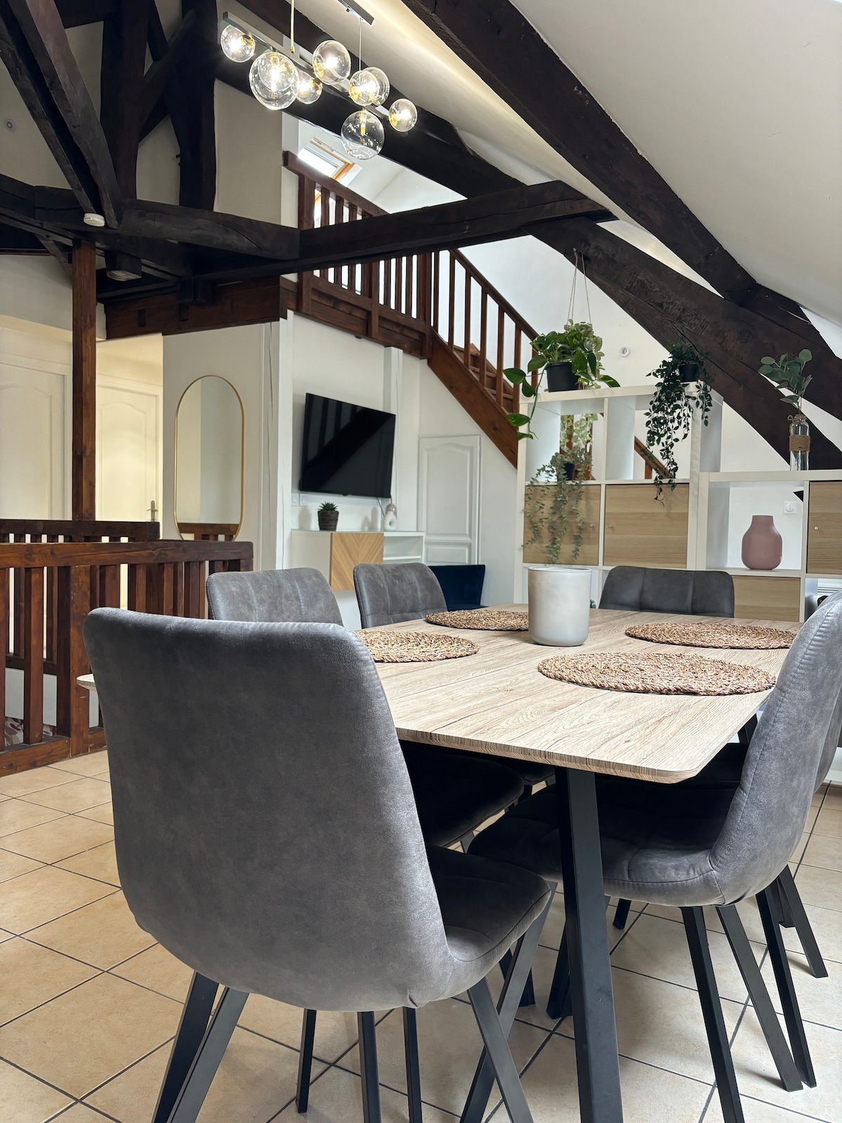 A dining area features a large wooden table surrounded by four dark grey chairs. Exposed wooden beams highlight the space, while a staircase leads to an upper level. A small plant sits on the table, and a TV is mounted on the wall in the background.