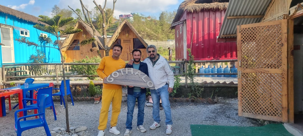 Three individuals are pictured in a vibrant outdoor setting, standing in front of uniquely designed huts. A mix of colorful chairs and a table are positioned nearby, and a decorative sign can be seen, contributing to the welcoming atmosphere.