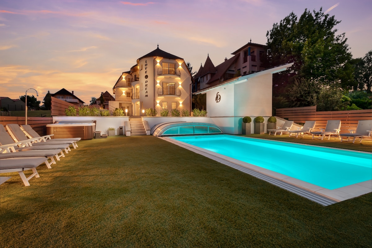 An inviting outdoor space features a well-lit pool with turquoise water surrounded by neatly arranged lounge chairs. The elegant building in the background showcases a modern design, with soft lighting accentuating its architectural details against a colorful sunset sky.