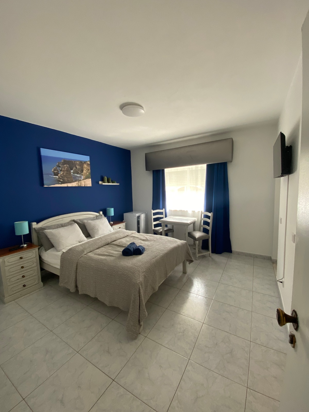 A serene bedroom features a spacious bed adorned with light bedding, complemented by a decorative throw. A small table with two chairs sits beside a window, with natural light filtering through sheer curtains. A television is mounted on the wall, enhancing the cozy setting.
