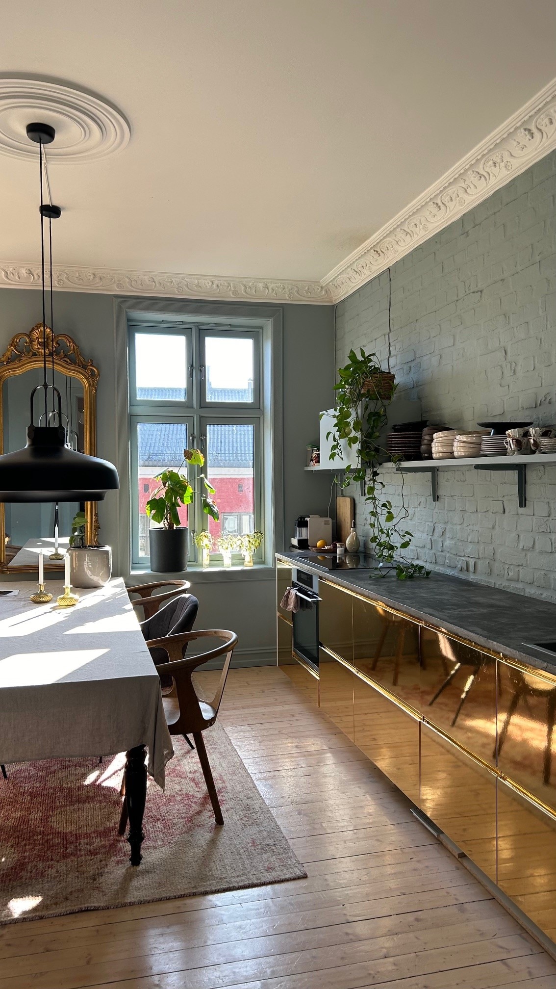 A well-lit kitchen showcases a large dining table draped with a light-colored tablecloth. Gold countertops reflect light and complement the greenery from potted plants. Shelves hold kitchenware and cookbooks, while a mirror and large window enhance the space's openness.