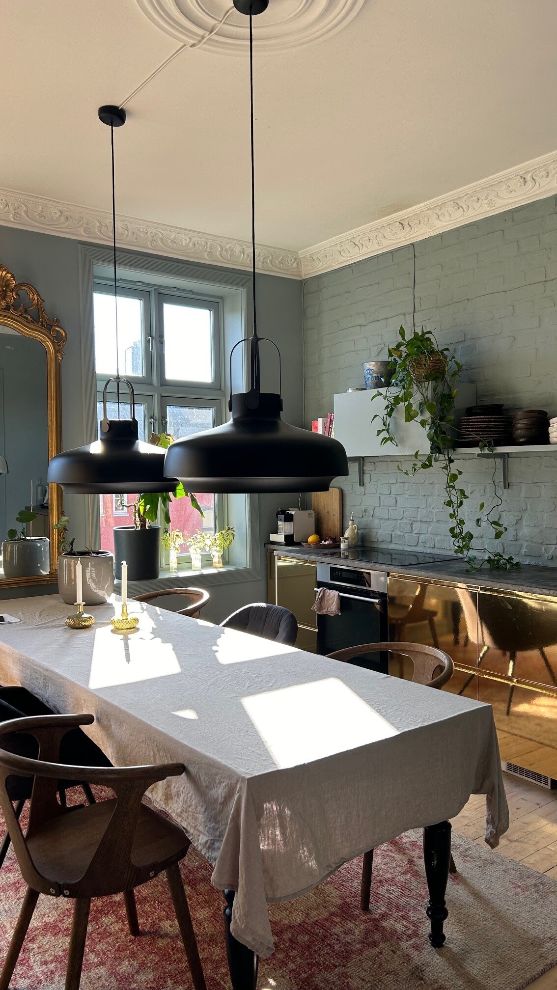 A dining area features a long table draped with a light-colored tablecloth, surrounded by wooden chairs. Two black pendant lights hang above the table, while natural light enters through large windows. A decorative mirror and greenery add touches of style to the space.