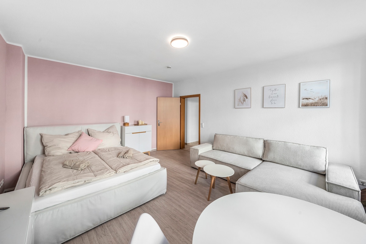 A spacious living area is presented, featuring a comfortable double bed with soft bedding and a decorative pillow. Nearby, a light-colored couch invites relaxation, accompanied by a round coffee table. Pale pink walls add a subtle touch, complemented by simple wall art.