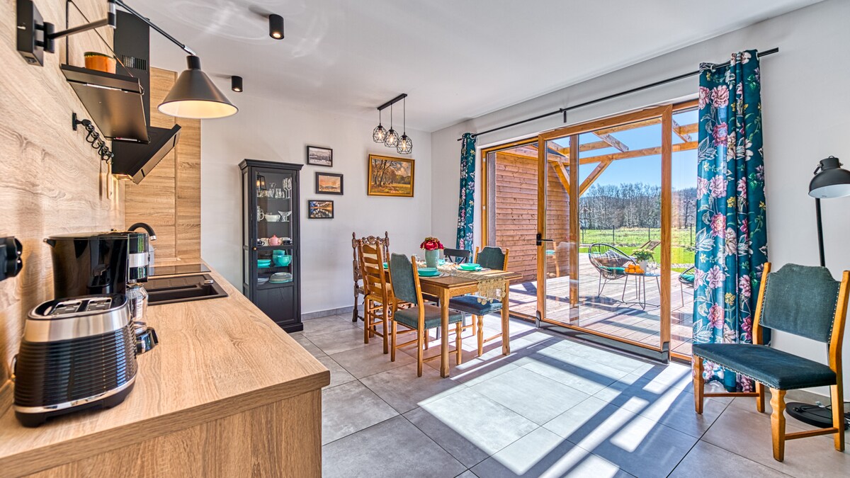 A modern kitchen and dining area is shown, featuring wooden cabinetry and a coffee machine. A dining table surrounded by six chairs is positioned near large sliding glass doors that open to a deck with outdoor seating, allowing natural light to fill the space.
