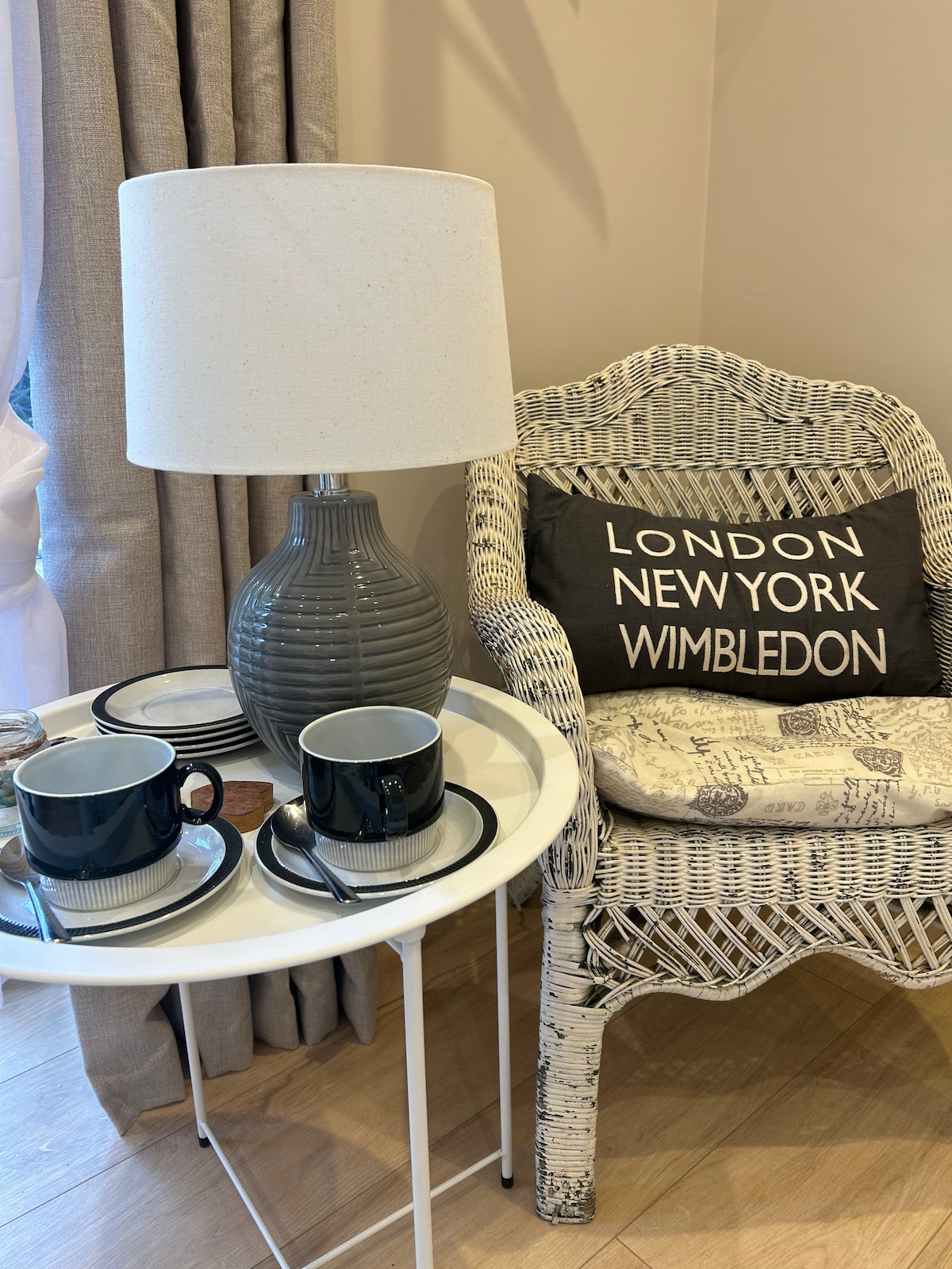 A cozy corner features a wicker chair adorned with a cushion displaying city names. A small round table holds two cups and saucers, accompanied by a textured lamp casting soft light nearby. Neutral curtains frame a window, enhancing the calming ambiance.