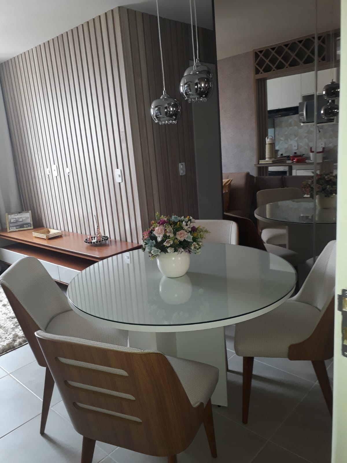 A modern dining area features a circular glass table surrounded by four upholstered chairs. A small vase of fresh flowers is centered on the table. The backdrop includes a decorative wooden wall and glimpses of the adjoining kitchen space.