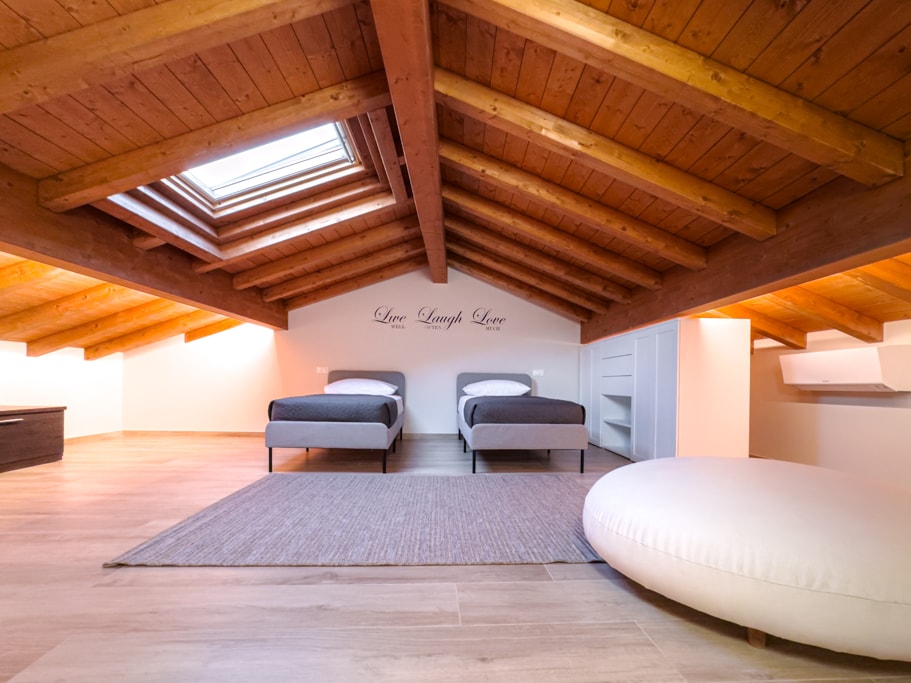 A spacious loft room features two beds with modern grey bedding arranged under a wooden-beamed ceiling. Natural light enters through a skylight, illuminating the light wooden flooring. A neutral rug and a circular ottoman provide additional comfort in the inviting space.