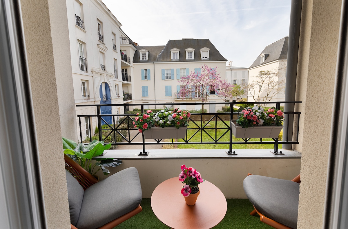 A serene balcony area is presented, featuring two comfortable chairs and a round table. Flower pots filled with blooms add color, while a soft green carpet enhances the tranquil setting. A view of the courtyard and surrounding buildings is visible, inviting relaxation.
