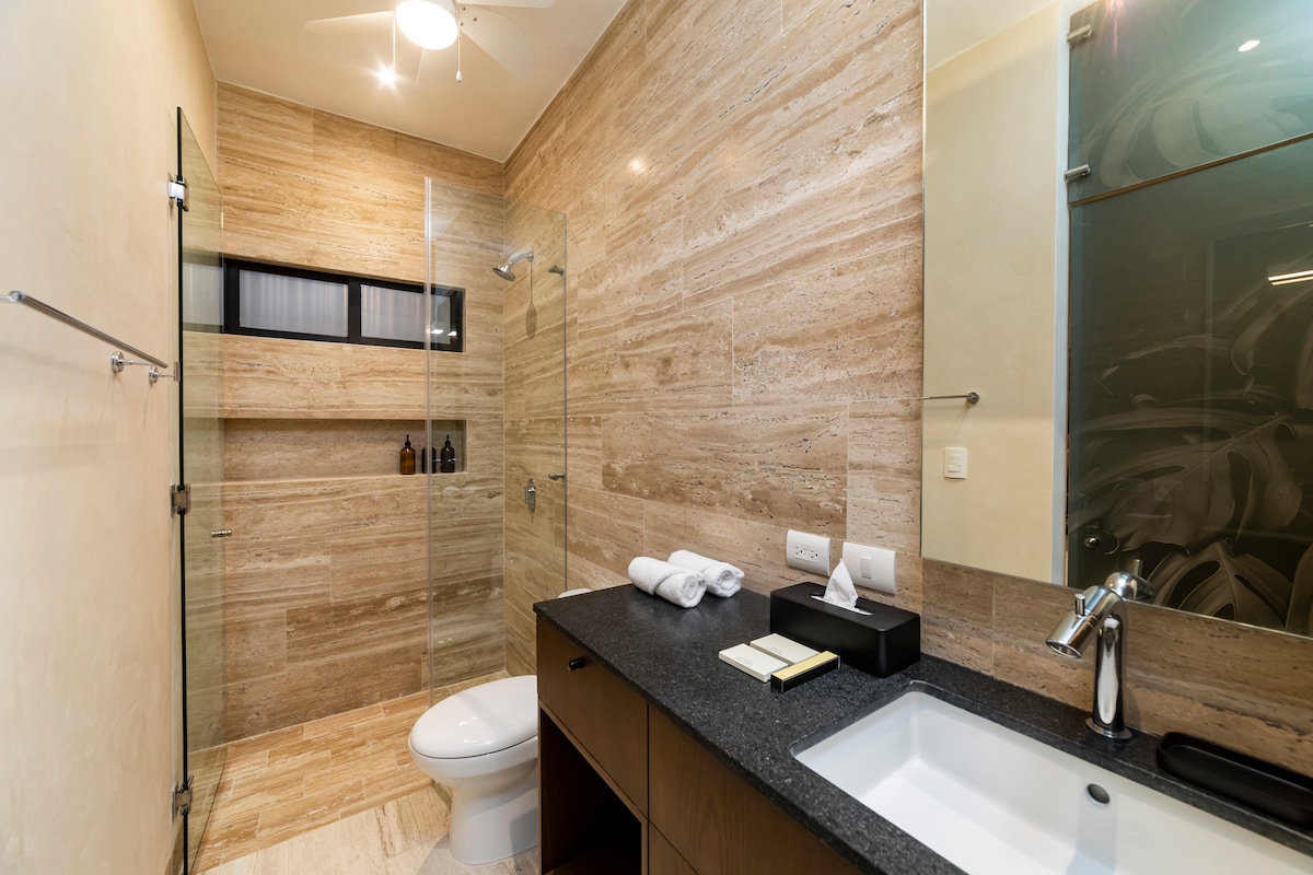 A modern bathroom features natural stone walls and a spacious walk-in shower. A sleek black countertop contrasts with light cabinetry. Toiletries are neatly arranged on a shelf, while a stylish sink adds to the minimalist aesthetic of the space.