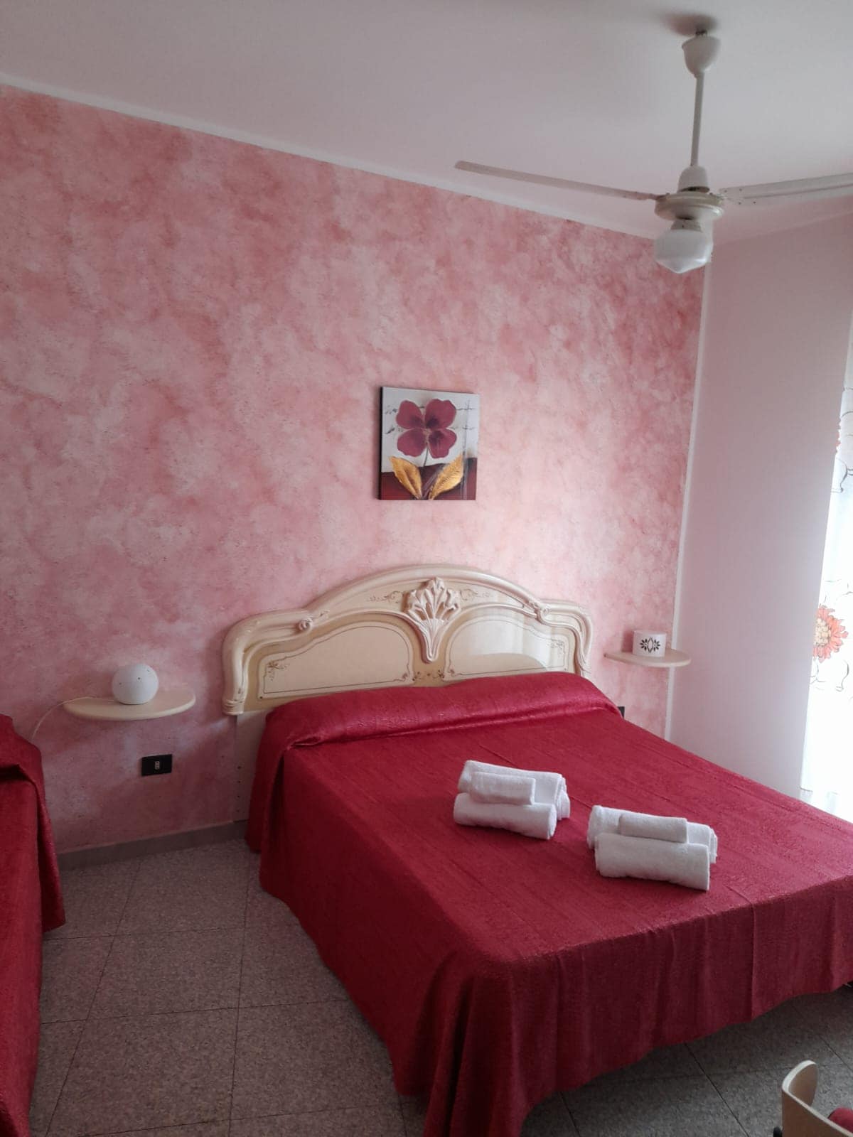 A private room features soft pink walls and two beds covered in red linens. A simple piece of wall art depicting a flower adds a decorative touch. Natural light enters through a nearby window, enhancing the room's inviting atmosphere.