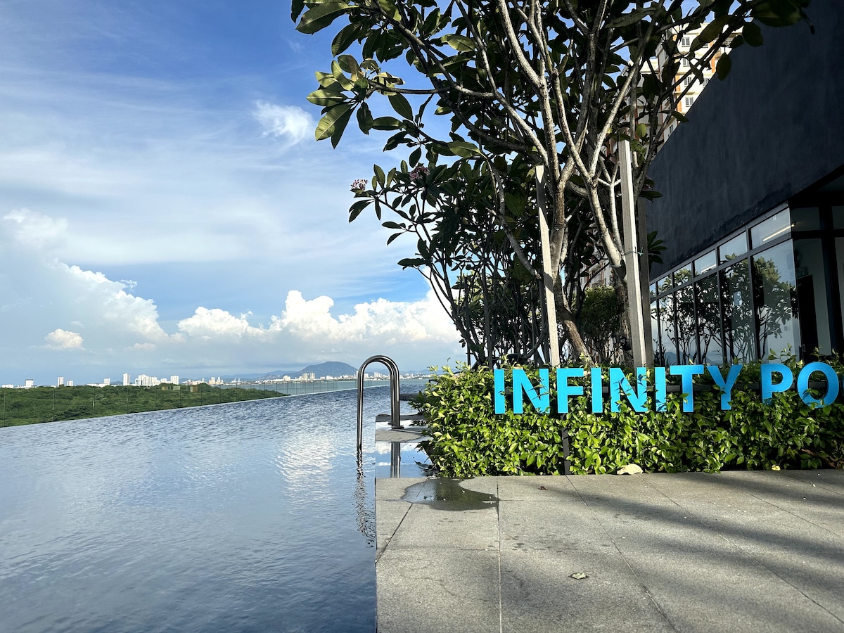 An infinity pool extends seamlessly towards the horizon, framed by lush greenery. The water reflects the sky, creating a tranquil atmosphere. A modern structure is visible nearby, enhancing the serene setting.