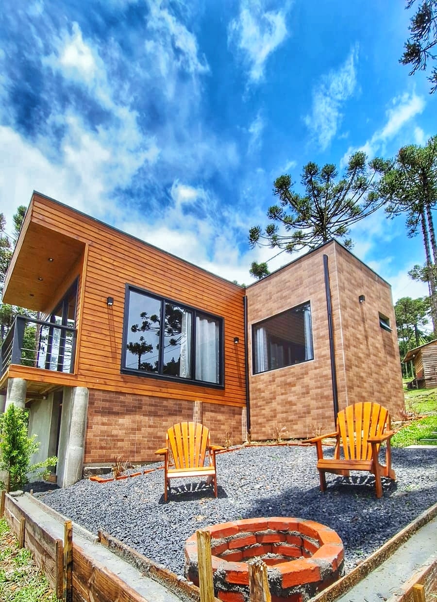 A modern chalet exterior is captured under a bright blue sky with scattered clouds. Two wooden chairs with a welcoming orange hue are positioned around a circular fire pit, set on a grassy area bordered by gravel. The building features large windows and natural wood accents.