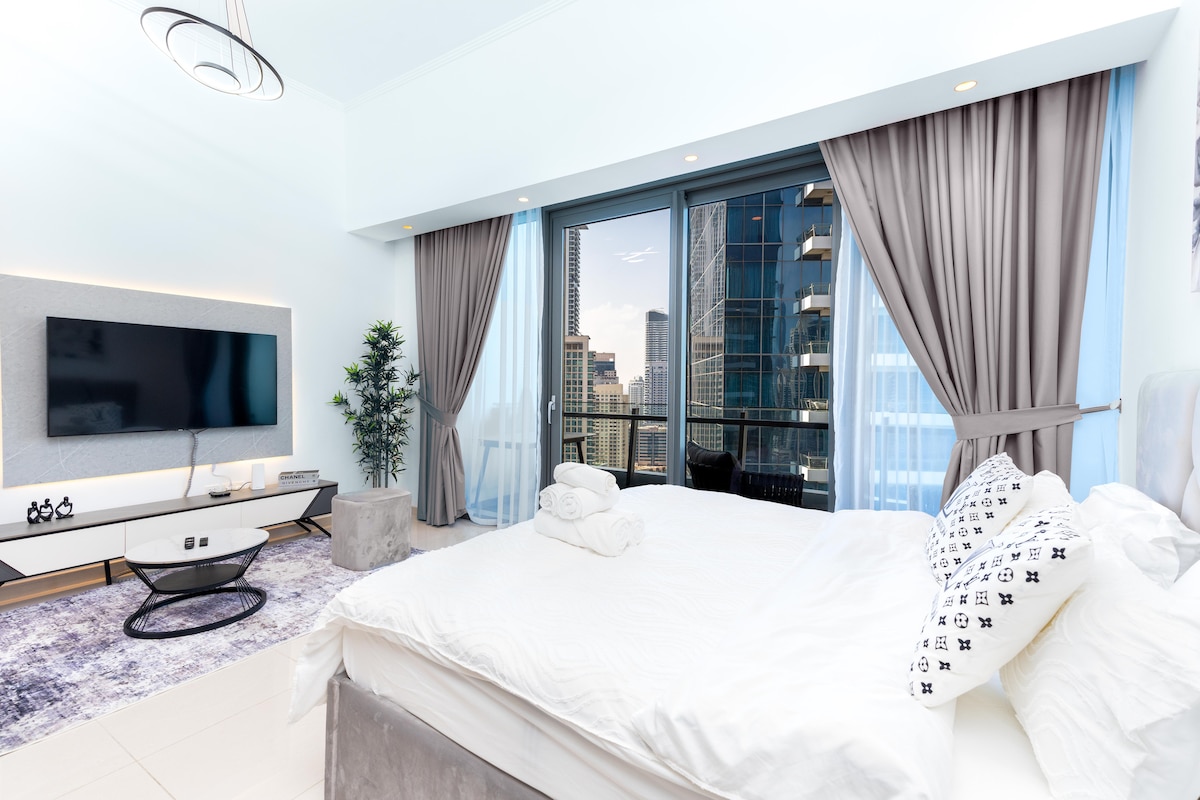 A serene bedroom is highlighted by a spacious bed adorned with crisp linens and decorative pillows. Large windows offer views of the city skyline, while sheer curtains gently diffuse natural light. A wall-mounted TV and minimalist decor contribute to a calm ambiance.