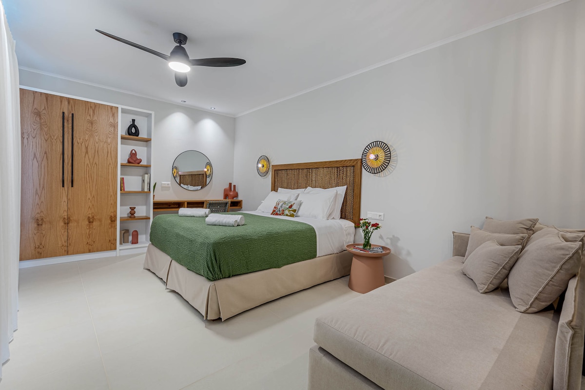 A serene sleeping area is presented, featuring a spacious bed with a green bedspread and decorative pillows. Natural light enters through a partially open curtain, reflecting off the light-colored walls. A cozy seating area is included, along with a stylish wooden wardrobe and shelves displaying decorative items.