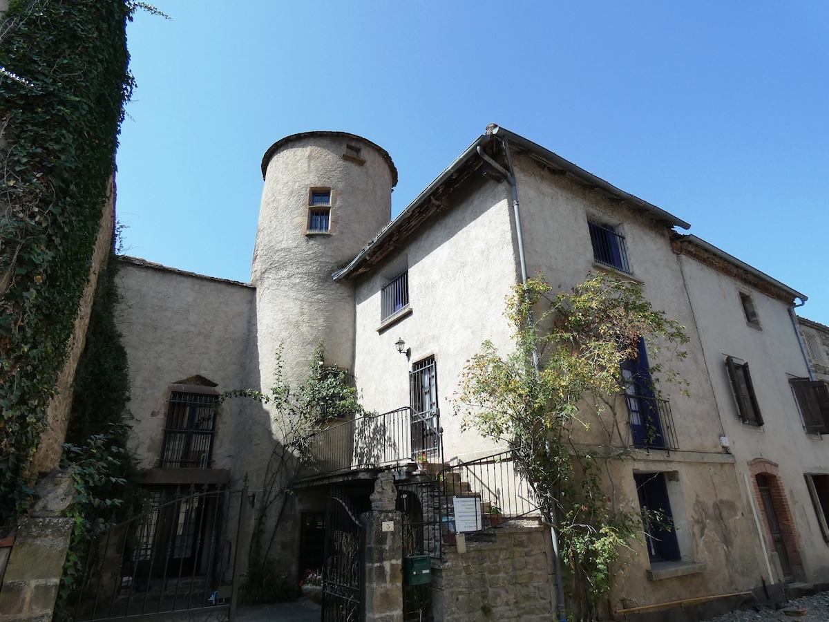 The exterior of a charming stone building is depicted, featuring a round tower and multiple windows. Lush greenery climbs the walls, while a small staircase leads to an entrance adorned with wrought iron railings. Clear blue skies complete the tranquil scene.