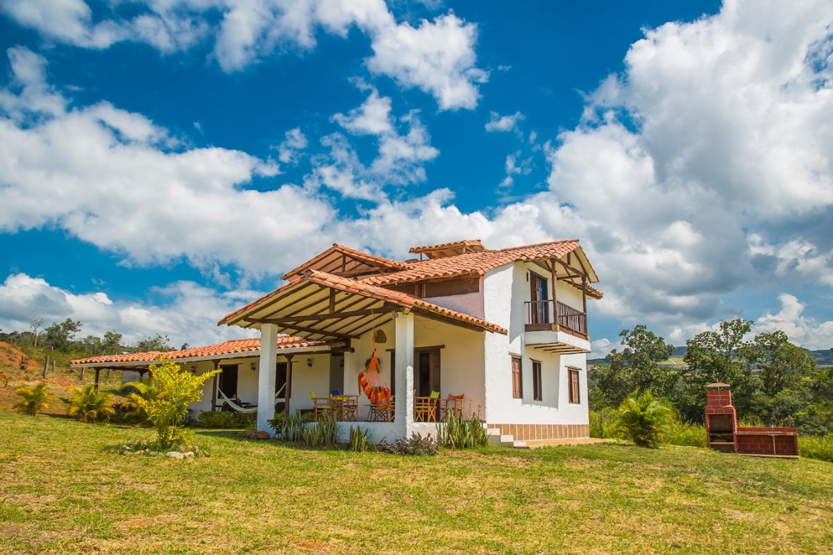 A two-story cabin is situated in a green landscape, featuring a traditional design with a terracotta tiled roof and whitewashed walls. Large windows and balconies provide views of the surrounding nature. Lush grass surrounds the home, enhancing the rural ambiance.