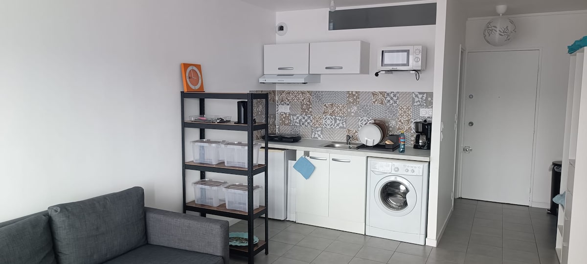 The kitchen area is equipped with modern appliances, including a microwave and a washing machine. A compact shelving unit holds clear storage containers. Decorative tiles accentuate the backsplash, while a small countertop workspace allows for meal preparation.