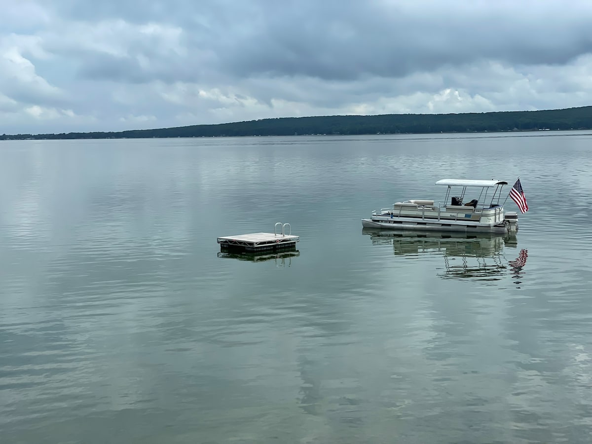 The calm surface of Hubbard Lake reflects a cloudy sky, creating a serene atmosphere. A swim raft and a pontoon boat are positioned on the water, with the boat displaying an American flag. The surrounding shoreline is softly visible in the background.