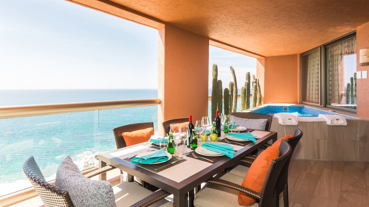 An outdoor dining area features a large table set with dishes and drinks, surrounded by comfortable chairs. The backdrop includes a scenic ocean view, with cacti visible in the distance. A small hot tub is situated nearby, adding to the relaxing ambiance.