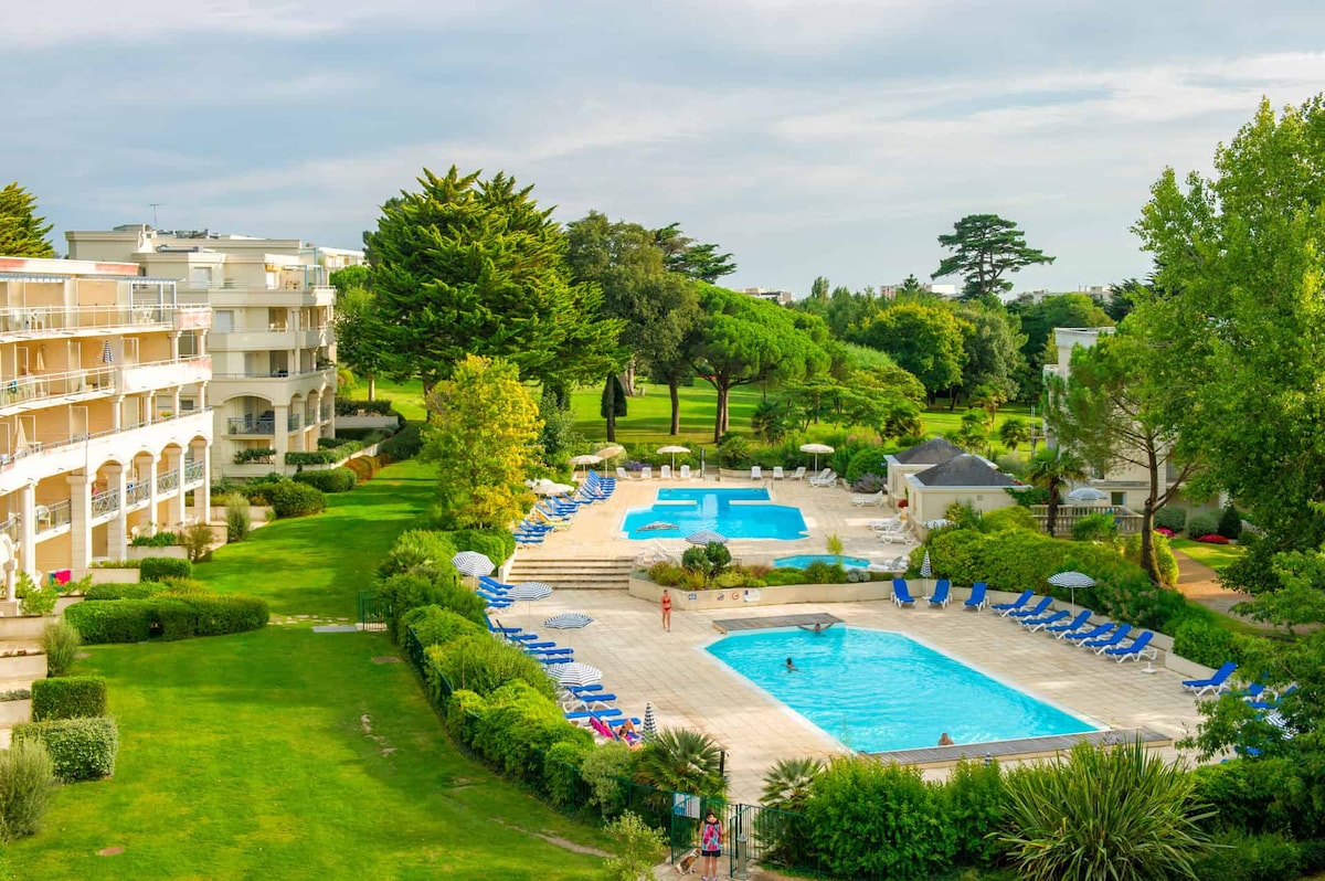 Two swimming pools are visible in the landscaped grounds of the Royal Park residence, surrounded by green lawns and trees. Sun loungers are arranged around the pool decks, with shades providing some relief from the sun. The area is spacious and well-maintained.