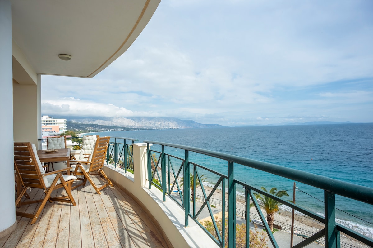 A spacious balcony offers stunning views of the sea, featuring wooden furniture including a table and chairs. The railing provides unobstructed sightlines to the shoreline and the distant mountains, while the calm waters reflect the sky.
