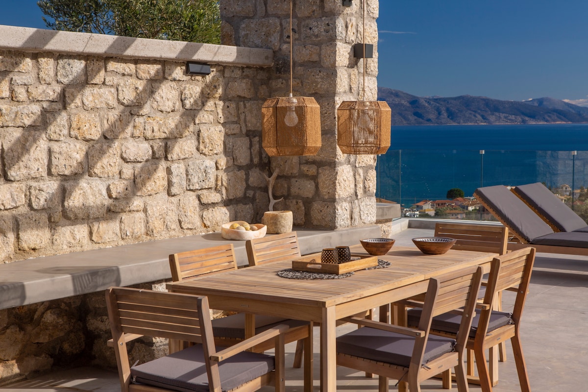 An outdoor dining area features a wooden table surrounded by matching chairs. Natural light casts shadows on the stone wall and smooth surfaces. Two pendant lights hang nearby, adding a warm touch. The stunning sea view and distant mountains can be seen in the background.