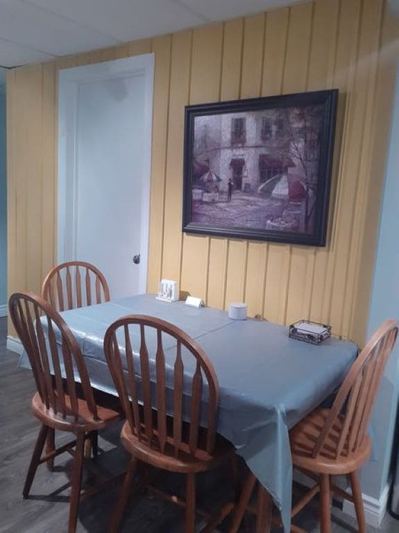 A dining area is presented with a wooden table surrounded by four matching chairs. A light blue tablecloth covers the table, complemented by a framed artwork depicting a serene street scene. Yellow paneled walls create a bright and welcoming backdrop for meals.
