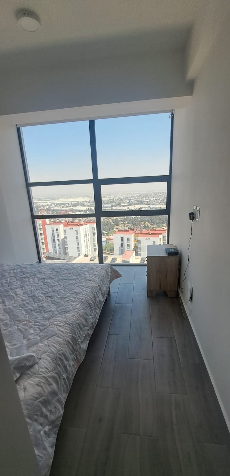 A well-lit sleeping area features a spacious bed with a textured white coverlet. A bedside table is positioned adjacent to the bed, and large windows provide a panoramic view of the surrounding landscape, enhancing the sense of openness and light.