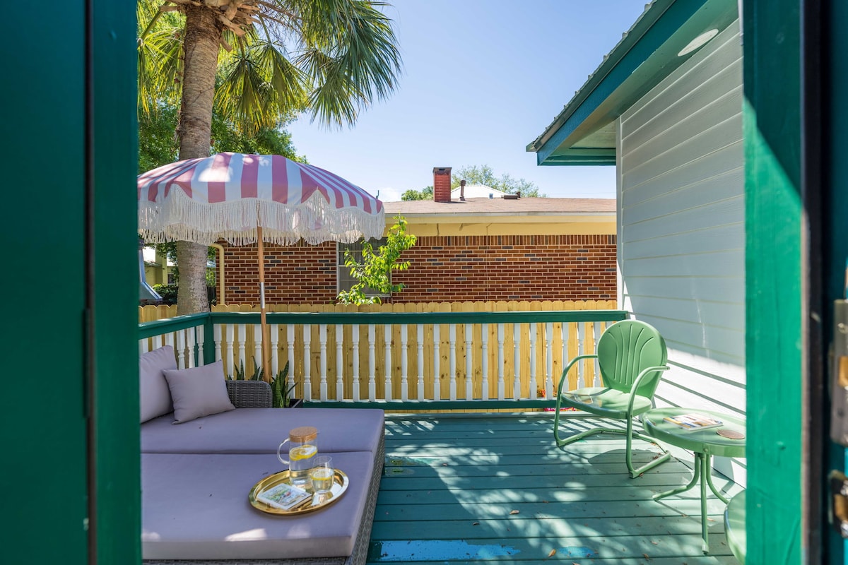 A charming outdoor space is visible, featuring a comfortable lounge area with a cushioned sofa in soft hues. A striped umbrella provides shade, while a green chair and small table complete the setting, with palm trees adding a tropical touch.