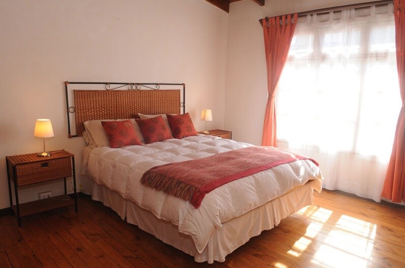 A spacious sleeping area features a large bed with a white comforter and decorative pillows. Two bedside tables with lamps are positioned beside the bed. Natural light floods the room through large windows dressed with sheer curtains, while warm wooden flooring complements the inviting setting.